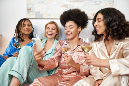 Fröhliche afrikanisch-amerikanische Frau hält verschwommenes Smartphone neben multiethnischen Freundinnen in bunten Pyjamas und hält Weingläser während der Nacht der Mädchen zu Hause, Bindungszeit, Schlummerparty