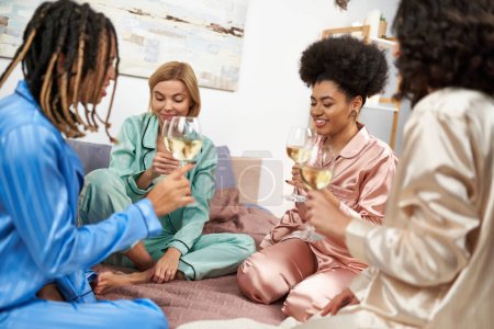 Femme afro-américaine souriante tenant un verre de vin tout en parlant à des copines multiethniques avec des verres de vin pendant la nuit des filles dans la chambre à coucher à la maison, liant le temps dans des vêtements de nuit confortables