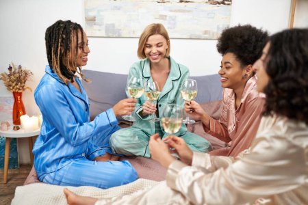 Foto de Novias positivas y multiétnicas en pijama colorido sosteniendo vasos de vino mientras habla durante la noche de las niñas en la cama en casa, tiempo de vinculación en ropa de dormir cómoda, fiesta de pijamas - Imagen libre de derechos