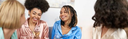 Sonrientes mujeres afroamericanas en pijama colorido sosteniendo copas de vino y hablando con novias borrosas durante la noche de las niñas en casa, tiempo de unión en ropa de dormir cómoda, pancarta 