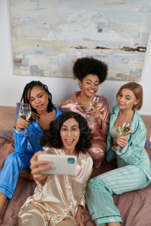 Foto de Vista de ángulo alto de alegres novias multiétnicas en pijamas coloridos sosteniendo vino y tomando selfie en el teléfono inteligente mientras está sentado en la cama en casa, tiempo de unión, fiesta de pijamas - Imagen libre de derechos