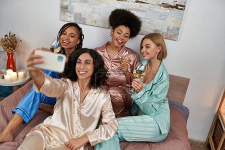 Téléchargez les photos : Vue en angle élevé de la femme multiraciale en pyjama prenant selfie avec des copines multiethniques gaies avec du vin et assis sur le lit à la maison, le temps de liaison dans des vêtements de nuit confortables, soirée pyjama - en image libre de droit