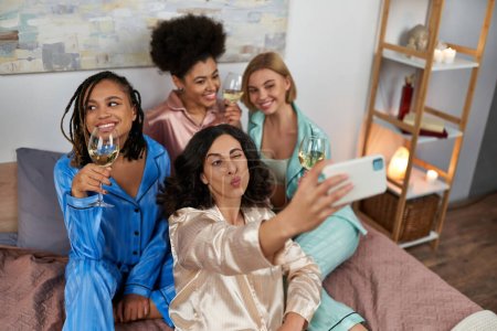 High angle view of multiracial woman blowing air kiss and taking selfie with multiethnic girlfriends with wine sitting on bed during pajama party at home, slumber party 