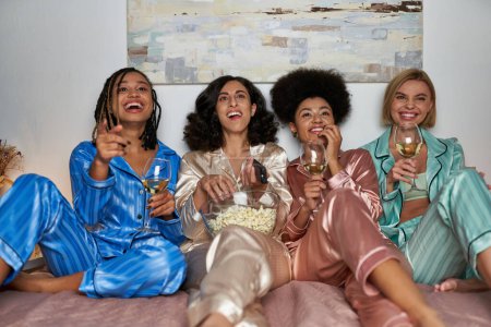 Cheerful multiethnic girlfriends in colorful pajama holding glasses of wine and popcorn while watching tv on bed during girls night, bonding time in comfortable sleepwear