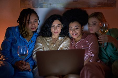 Smiling multiethnic girlfriends in colorful pajama holding glasses of wine and using blurred laptop during girls night at home, bonding time in comfortable sleepwear, slumber party