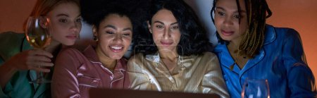 Mujeres alegres y multiétnicas en pijama colorido usando una computadora portátil borrosa y sosteniendo copas de vino durante la noche de las niñas en casa, tiempo de unión en ropa de dormir cómoda, pancarta 
