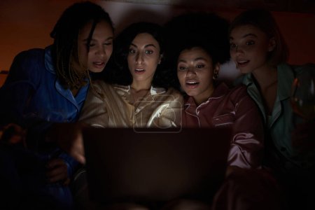 Novia multicultural conmocionada en pijama colorido mirando juntos en la computadora portátil borrosa durante la fiesta de pijama en la noche en casa, tiempo de unión en ropa de dormir cómoda, película de miedo 