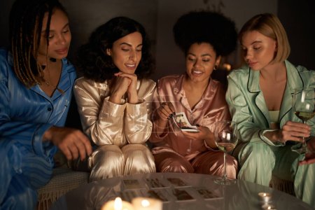 Lächelnde Afroamerikanerin mit Tarot-Karten neben multiethnischen Freundinnen mit Wein und Kerzen bei Pyjama-Party zu Hause, Bindungszeit in bequemer Nachtwäsche, Wahrsagerei 