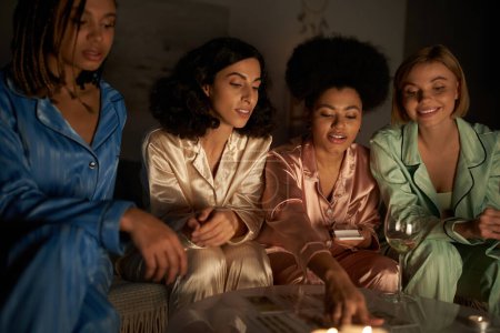 Foto de Sonrientes novias multiétnicas en pijama colorido mirando las cartas del tarot en la mesa cerca de un vaso de vino y velas durante la noche de las niñas en casa, tiempo de unión en ropa de dormir cómoda, fiesta de pijamas - Imagen libre de derechos