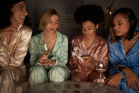Positive multiethnische Freundinnen im Pyjama sitzen neben afrikanisch-amerikanischen Freundinnen mit Tarot-Karten und Weingläsern während der Nacht der Mädchen zu Hause, Bindungszeit in bequemer Nachtwäsche, Weissagung 