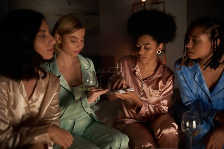 Young woman in pajama holding glass of wine and taking tarot card near multiethnic girlfriends during girls night at home, bonding time in comfortable sleepwear, divination 