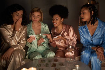 Novias multiétnicas positivas en pijama colorido sosteniendo cartas del tarot y hablando cerca de copas de vino y velas durante la fiesta de pijama en casa, tiempo de unión en ropa de dormir cómoda
