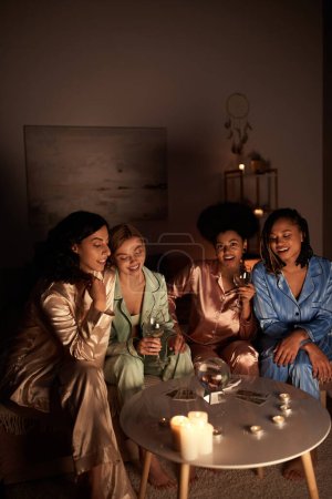 Positive multiethnische Freundinnen im farbenfrohen Pyjama mit Weingläsern in der Nähe von Tarotkarten, Kristallkugel und Kerzen während der Nacht der Mädchen zu Hause, Bindungszeit in bequemer Nachtwäsche, Wahrsagerei 