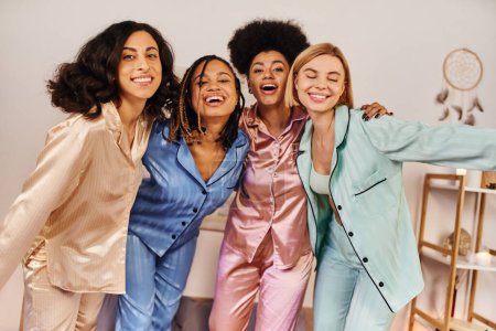 Lächelnde multiethnische Freundinnen im farbenfrohen Pyjama, die in die Kamera schauen, einander umarmen und bei der Schlummerparty zu Hause Spaß haben, Zeit in bequemer Nachtwäsche verbinden