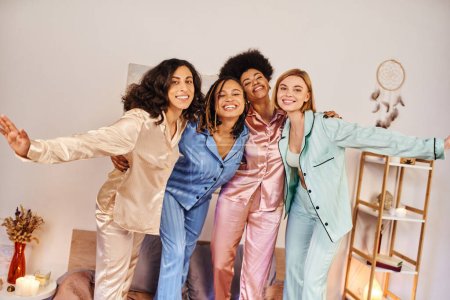 Lächelnde multikulturelle Freundinnen im farbenfrohen Pyjama, die sich umarmen und einander anschauen, während sie zu Hause auf dem Bett stehen und in bequemer Nachtwäsche Zeit miteinander verbringen