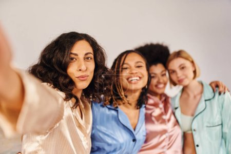 Multiracial woman in pajama looking at camera while standing near cheerful and blurred multiethnic girlfriends at home during slumber party, bonding time in comfortable sleepwear