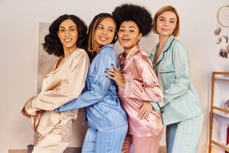 Fröhliche multikulturelle Freundinnen im bunten Pyjama, die in die Kamera schauen und sich beim Schlummerfest zu Hause umarmen, kulturelle Vielfalt, Bindungszeit in bequemer Nachtwäsche