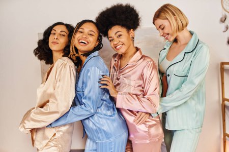 Glückliche multikulturelle Freundinnen im bunten Pyjama, die in die Kamera schauen, Spaß haben und sich bei Pyjama-Party zu Hause umarmen, Zeit in bequemer Nachtwäsche verbringen, kulturelle Vielfalt