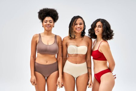 Smiling multiethnic women in colorful lingerie looking at camera together and standing isolated on grey, different body types and self-acceptance, multicultural representation, cultural diversity