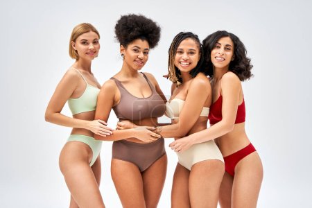 Cheerful multicultural girlfriends in colorful and modern lingerie hugging each other and standing isolated on grey, different body types and self-acceptance, multicultural representation