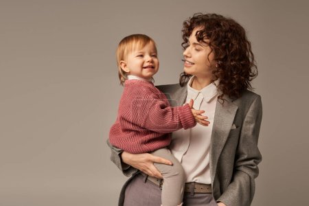 work life balance concept, happy woman in suit holding daughter and smiling on grey background, career and family, loving motherhood, quality time, successful mother 