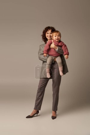 Photo for Quality time, career and family, happy woman in suit holding child on grey background, growth and family, loving motherhood, lifestyle, multitasking, work life harmony concept - Royalty Free Image