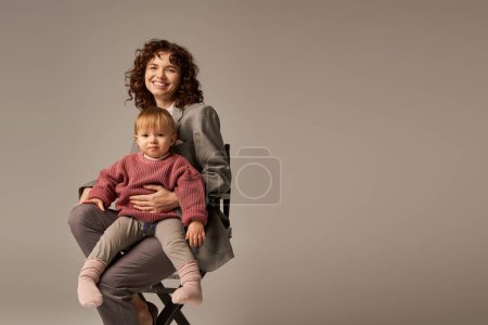 balanced lifestyle, career and family, curly businesswoman sitting on chair with child on grey background, professional success, motherhood, multitasking, quality time, work life balance concept 
