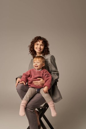 balanced lifestyle, growth and family, happy businesswoman sitting on chair with happy child on grey background, professional success, motherhood, quality time, work life balance concept 