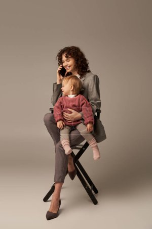 mode de vie équilibré, appel téléphonique, femme d'affaires heureuse parlant sur smartphone et assise sur une chaise avec enfant sur fond gris, maternité, multitâche, temps de qualité, concept d'équilibre entre vie professionnelle et vie privée 