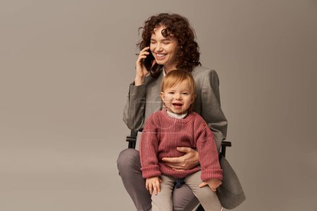 balanced lifestyle, phone call, happy working mother talking on smartphone and sitting on chair with child on grey background, motherhood, multitasking, quality time, work life balance concept 