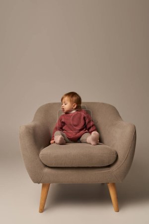 niedliche Baby-Mädchen, Kleinkind in Freizeitkleidung auf bequemen Sessel auf grauem Hintergrund im Studio sitzen, Emotionen, verwirrt, Unschuld, kleines Kind, Kleinkind Mode, stilvolles Outfit, Pullover 