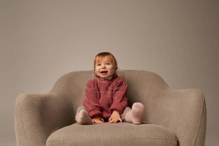 cute baby girl, toddler in casual attire sitting on comfortable armchair on grey background in studio, emotion, happiness, joy, innocence, little child, toddler fashion, stylish outfit, sweater 