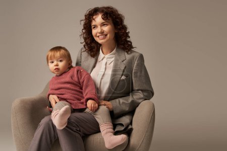 modern working parent, curly woman in suit sitting in armchair with toddler daughter, grey background, happy mother and child, multitasking, quality family time, balancing work and life concept 