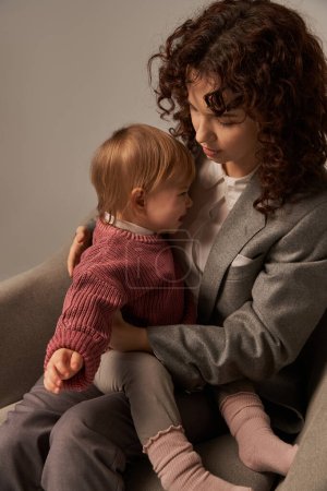 moderne berufstätige Eltern, Balanceakt zwischen Arbeit und Lebenskonzept, lockige Frau im Anzug im Sessel sitzend mit Kleinkind-Tochter, beruhigendes weinendes Baby, grauer Hintergrund, Mutter und Kind, Multitasking 