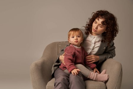 balancing work and life concept, curly woman in suit sitting in armchair with toddler daughter, grey background, multitasking, quality family time, mother and child, modern working parent 