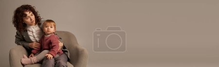 Foto de Equilibrio entre el trabajo y el concepto de vida, mujer rizada en traje sentado en sillón con hija pequeña, fondo gris, multitarea, tiempo de familia de calidad, madre e hijo, padre trabajador moderno, pancarta - Imagen libre de derechos