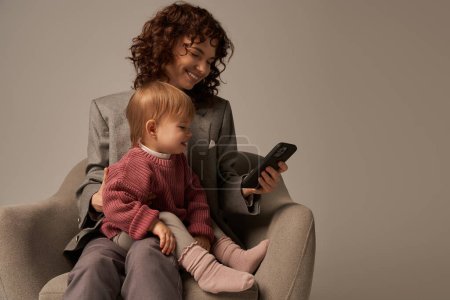 modern working parent, balancing work and life concept, happy woman in suit sitting with toddler daughter, using smartphone, grey background, happy mother and child, multitasking 