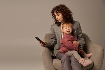 Multitasking, Smartphone-Nutzung, moderne berufstätige Mutter, Spagat zwischen Arbeit und Lebenskonzept, lockige Frau im Anzug mit Kleinkind auf Sessel sitzend, grauer Hintergrund, Eltern und Kind  