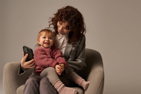 Photo for Modern working mother, multitasking, using smartphone, balancing work and life concept, happy businesswoman in suit sitting with toddler child on armchair, grey background, parent and child - Royalty Free Image
