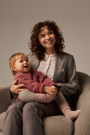 modern working mother, balancing work and life concept, happy businesswoman in suit sitting with toddler daughter on armchair, engaging with child, grey background, family relationships 