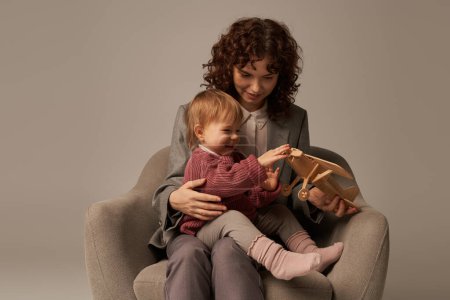 modern working mother, balancing work and life concept, businesswoman in suit sitting on armchair and playing with toddler daughter, wooden biplane, grey background, engaging with child 