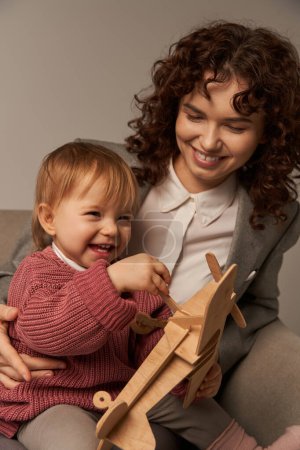 successful mother, balancing work and life concept, happy businesswoman in suit sitting on armchair and playing with toddler daughter, wooden biplane, grey background, engaging with child 