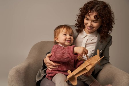 career and family, balancing work and life concept, businesswoman in suit sitting on armchair and playing with toddler daughter, wooden biplane, grey background, engaging with child 