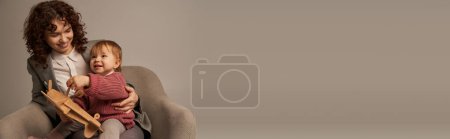 Téléchargez les photos : Mère de travail moderne, équilibre travail et concept de vie, femme d'affaires en costume assis sur un fauteuil et jouer avec la fille tout-petit, biplan en bois, fond gris, engagement avec l'enfant, bannière - en image libre de droit