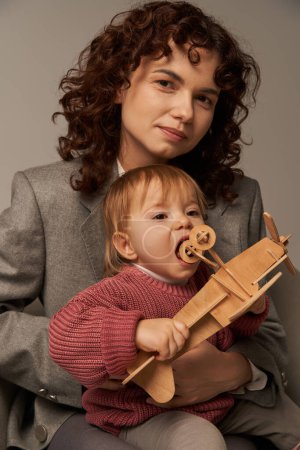 moderne berufstätige Mutter, Spagat zwischen Arbeit und Lebenskonzept, Geschäftsfrau im Anzug sitzt auf Sessel mit Kleinkind-Tochter, spielt mit hölzernem Doppeldecker, grauer Hintergrund, Mutterschaft 