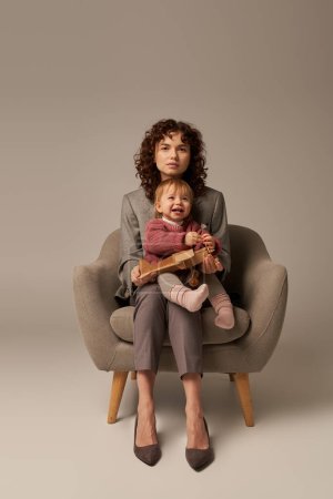 career and family, balancing work and life concept, businesswoman in suit sitting on armchair with toddler daughter, playing with wooden biplane, grey background, motherhood, full length 