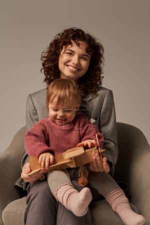 modern working parent, balancing work and life concept, businesswoman in suit sitting on armchair and playing with toddler daughter, wooden biplane, grey background, engaging with child 