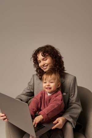 Photo for Building successful career, balanced lifestyle, working parent, happy curly woman using laptop and sitting on armchair with daughter, mother and child, motherhood, multitasking - Royalty Free Image