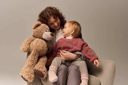 Photo for Quality family time, parenting and career, happy businesswoman holding teddy bear near toddler daughter on grey background, sitting on armchair, work life harmony concept, working parent - Royalty Free Image