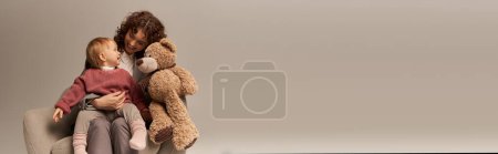 Qualität Familienzeit, Elternschaft und Karriere, lockige Geschäftsfrau hält Teddybär in der Nähe von Kleinkind Tochter auf grauem Hintergrund, sitzt auf Sessel, Work Life Harmony Konzept, berufstätige Eltern, Banner 